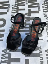 Sandalias de tacón alto Mutma modelo Ñoma, confeccionadas completamente en terciopelo color verde militar. Presentan una plataforma delantera, detalle de doble nudo o moña en la puntera, taco ancho y tira con hebilla ajustable en el tobillo.