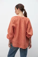 Camisa de corte holgado en color ocre, con escote en V y cordones con borlas decorativas. Presenta mangas largas abullonadas con puños elásticos y un diseño de textura rústica.