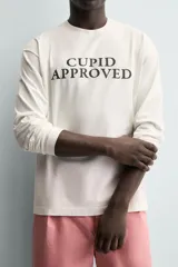 Camiseta de manga larga color blanco roto, corte relajado (relaxed fit) con cuello redondo y texto estampado en negro en el pecho que dice "CUPID APPROVED".