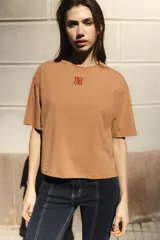 Remera de manga corta color camel, con cuello redondo y corte boxy. Presenta un pequeño logo bordado en hilo rojo en el pecho.