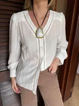 Camisa blanca de tela liviana con textura sutil, presenta detalles calados verticales en el delantero y volados delicados en los hombros. Posee mangas largas con puños y cierre frontal mediante botones.