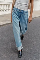Jeans baggy de tiro medio con pernera holgada, diseño de cinco bolsillos y bajo acabado en vuelta. Cuenta con cintura elástica en la espalda y cierre frontal con cremallera y botón.