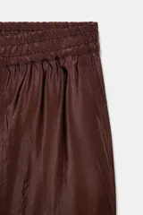 Pantalón ancho de tiro alto, color marrón oscuro, con cintura elástica fruncida y bolsillos delanteros. El tejido presenta un acabado satinado y arrugado.