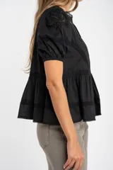 Blusa negra de algodón con corte oversize, mangas cortas abullonadas y detalles de encaje y bordados en el frente y hombros. Presenta un diseño con pliegues verticales y terminaciones con puntilla.
