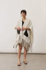 Ruana de punto color beige con detalles geométricos negros y flecos largos tricolores en el borde inferior.
