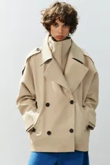 Trench corto oversize color beige con cuello subido de efecto doble, cierre cruzado con botones oscuros y bolsillos de vivo delanteros.