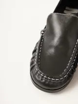 Mocasines de cuero negro con costuras pespunteadas en hilo blanco a contraste, diseño fruncido en la puntera, talón plano con pequeño tacón y apliques sutiles en charol.