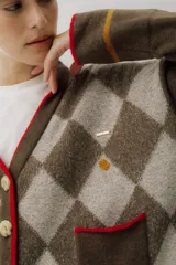 Saco tipo cardigan de punto con diseño de rombos en tonos marrón chocolate y beige. Presenta cierre frontal con botones grandes, cuello en V con borde en color rojo, dos bolsillos delanteros con ribete rojo y detalles bordados pequeños. Posee mangas largas con una franja horizontal en color mostaza cerca del puño.