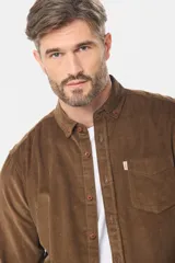 Camisa de pana fina en color verde oscuro, de manga larga con puños arremangados, cuello camisero con botones y un bolsillo aplicado en el pecho con pequeña etiqueta decorativa.