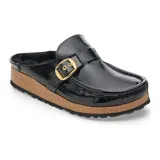 Zueco tipo mule de cuero charol negro con interior forrado en piel sintética. Presenta una hebilla metálica dorada ajustable en el empeine y una suela de corcho con base dentada de goma.