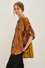 Camisa de manga corta con cuello mao y calce amplio, confeccionada en viscosa con estampado tie-dye en tonos ocre, negro y violeta. Presenta detalles bordados con mostacillas, lentejuelas, canutillos e hilos de lurex, y cierre frontal con botones a tono.