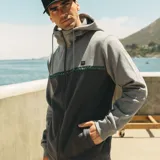 Campera canguro Maui and Sons de felpa con cierre completo y capucha ajustable con cordón. Presenta un diseño bicolor en tonos gris claro y gris oscuro, separado por una franja horizontal con patrón de olas y detalles en turquesa. Cuenta con bolsillos laterales tipo ojal y puños y ruedo de rib elástico.