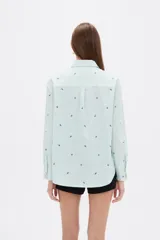 Blusa de corte holgado con cuello camisero y cierre frontal de botones. Presenta un diseño de rayas verticales finas en color azul y blanco, decorado con pequeños bordados dispersos.