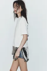 Camiseta negra de corte oversize, cuello redondo y manga corta. Presenta un bajo asimétrico con una aplicación de lentejuelas plateadas a contraste.