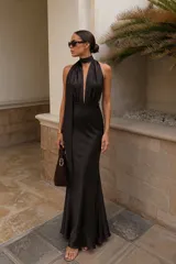 Vestido largo de satén negro con caída fluida, escote profundo tipo halter con tiras para anudar al cuello y diseño de espalda descubierta.