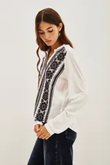 Camisa de algodón y viscosa color blanco, con mangas largas y diseño frontal con bordados geométricos y florales en contraste negro. Presenta cuello redondo con pequeña abertura y cierre frontal de botones.