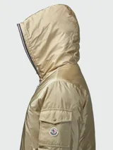 Campera tipo plumífero corto de Moncler, color beige con acabado satinado. Presenta capucha integrada, cierre frontal con cremallera, bolsillos laterales y un bolsillo con solapa en la manga izquierda que incluye el logo de la marca.