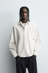 Camisa de corte relajado confeccionada en tejido de algodón, con diseño de rayas verticales. Presenta cuello solapa, cierre frontal de botones, manga larga con puños abotonados y un bolsillo plastrón en el pecho con un pequeño bordado decorativo.
