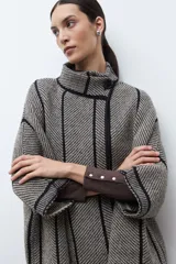 Capa de tejido de punto en color beige y negro con patrón de espiga, cuello alto y cierre de botón. Presenta vivos negros en contraste que delinean el diseño.