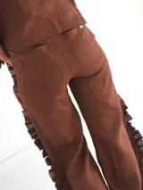 Pantalón de lino color beige, con apliques de tela con volados y costuras decorativas que aportan textura y movimiento. Cuenta con diseño de pernera ancha, bolsillos y aberturas laterales.