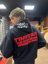 Hoodie negro de corte oversize con capucha, confeccionado en algodón. Presenta un estampado gráfico en color rojo en la espalda con la leyenda 'ITTAI STARS THE STAR BRAND DESIGNED IN VENICE' y un detalle bordado en blanco en la parte posterior de la capucha.