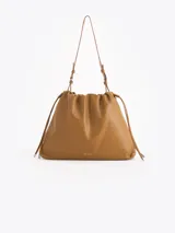 Cartera tipo shopper de cuero vacuno color tostado, con formato amplio, fruncido superior y cordones ajustables a los lados. Cuenta con tiras finas para llevar al hombro y sutil grabado de la marca en el frente.