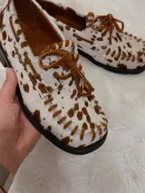 Zapato tipo mocasín de cuero con pelo estampado con diseño de manchas de vaca en tonos marrón y blanco. Presenta cordones frontales, costuras visibles en el borde y suela plana.