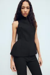 Chaleco entallado de color negro con cuello alto y cierre frontal con cremallera oculta. Presenta un diseño sin mangas con costuras verticales que definen la silueta y un bajo con ligero volumen acampanado.