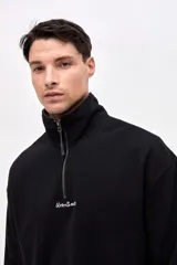 Buzo de felpa con friza, color negro, con cuello alto y cierre frontal parcial. Presenta bordado de la marca en el pecho y un calce relajado.