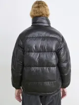 Campera puffer de corte holgado en color marrón oscuro con acabado brillante. Presenta cuello alto, cierre frontal con cremallera y botones a presión, además de bolsillos frontales con solapa.