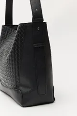 Cartera de hombro color negro con diseño trenzado en el panel frontal y laterales lisos. Cuenta con una correa larga ajustable y una placa metálica pequeña con el logo de la marca en la parte inferior.