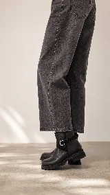 Bota corta de cuero negro con suela track de goma, detalle de tira lateral con hebilla metálica y acabado minimalista.