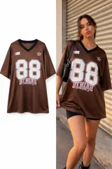 Remerón de estilo deportivo en color marrón, con cuello en V negro y mangas cortas. Presenta el número 88 estampado en blanco con bordes rosados en el frente, junto con detalles gráficos adicionales en la parte superior y texto en la zona inferior.