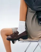 Mocasín de cuero marrón con textura granulada, diseño de horma cuadrada, detalle de flecos y lazo decorativo en el empeine, montado sobre una suela y taco bajo de color negro.