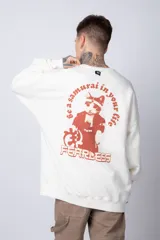 Buzo de tejido friza color crema, de corte oversize y cuello redondo. Presenta un pequeño bordado o parche en el pecho izquierdo con un diseño de cabeza de animal en tonos rojizos/terracota.