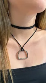 Gargantilla doble de gamuza negra que combina un choker ajustado al cuello y un collar largo con un dije metálico en forma de estribo.