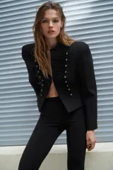 Blazer corta de estilo militar en color negro, con cuello redondo, manga larga y hombreras marcadas. Presenta un diseño frontal con bordados decorativos a tono y botones dorados, cierre con gancho metálico y forro interior.