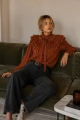 Camisa de color terracota con cuello alto, cierre frontal con lazo ajustable y volados decorativos en el pecho y hombros. Presenta mangas largas con puños elásticos.