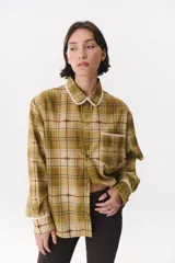 Camisa de corte holgado con estampado de cuadros en tonos mostaza, marrón y azul. Presenta cuello clásico, cierre frontal con botones, bolsillo en el pecho y detalles de puntilla decorativa en el cuello, bolsillo y puños.