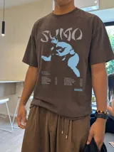 Remera marrón oversize con efecto acid wash, corte amplio y mangas cortas. Presenta un estampado frontal en tonos celestes y grises con la inscripción 'Sumo', una ilustración de un luchador de sumo y detalles tipográficos alusivos a un álbum musical.
