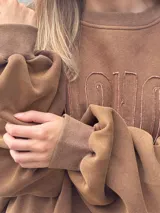 Buzo oversize color marrón con cuello redondo, mangas largas amplias y ruedo desflecado. Presenta letras en relieve con efecto de costura en el frente.