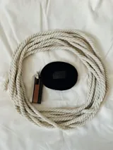 Monedero circular de color negro con cierre perimetral. Presenta una etiqueta frontal con el logo de la marca y una correa lateral de tela con franjas en tonos beige, marrón y negro.