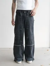 Pantalon baggy-flared de denim negro con efecto gastado, diseño desmontable con cierres horizontales en las piernas. Presenta múltiples parches de refuerzo, apliques metálicos, ojales decorativos y correas ajustables en los tobillos. Incluye un cinturón textil con tachas.