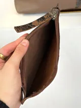Cartera tipo hobo de cuero genuino con diseño bicolor que combina paneles de cuero liso marrón con paneles de cuero texturizado con efecto desgastado en tonos negro y marrón. Incluye un sobre extraíble a juego con cierre y etiqueta de la marca.