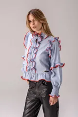 Camisa de algodón color celeste con volados decorativos en el frente, hombros y mangas, resaltados con un borde en color naranja. Presenta cierre frontal con botones, cuello alto con volado y mangas largas con puños ajustados.