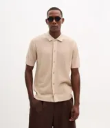 Camisa de punto de manga corta en color beige, con cuello inglés y cierre frontal de botones. Presenta una textura tejida y un corte regular que ofrece un ajuste cómodo y clásico.