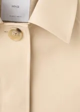 Chaqueta corta de algodón color beige, de corte holgado, con cuello clásico, cierre frontal de botones y bolsillos ribeteados. Cuenta con mangas largas amplias con puños ajustables mediante tiras y botones.