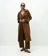 Trench coat largo de gamuza sintética con cuello peaked, cierre frontal cruzado con botones y cinturón ajustable en la cintura. Presenta mangas largas con reguladores en los puños y forro interior.