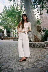 Top corto asimétrico color blanco roto, con un solo hombro descubierto y manga abullonada corta en el otro lado. Presenta bordados perforados (ojales) y un volante en el hombro descubierto. El bajo y el puño de la manga terminan en elástico.