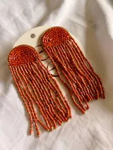 Aros colgantes de estilo artesanal, confeccionados con cuentas tubulares en color naranja y detalles en dorado. Presentan un diseño de semicírculo superior con flecos largos que caen en cascada.